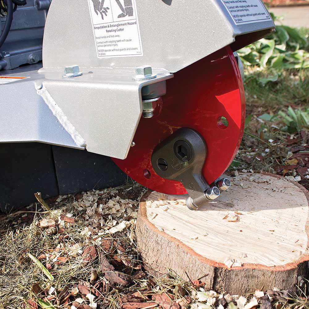 A close-up view of the Dosko 200-6HC Mini Stump Grinder's cutting wheel, which has a prominent warning label, as it grinds a tree stump.