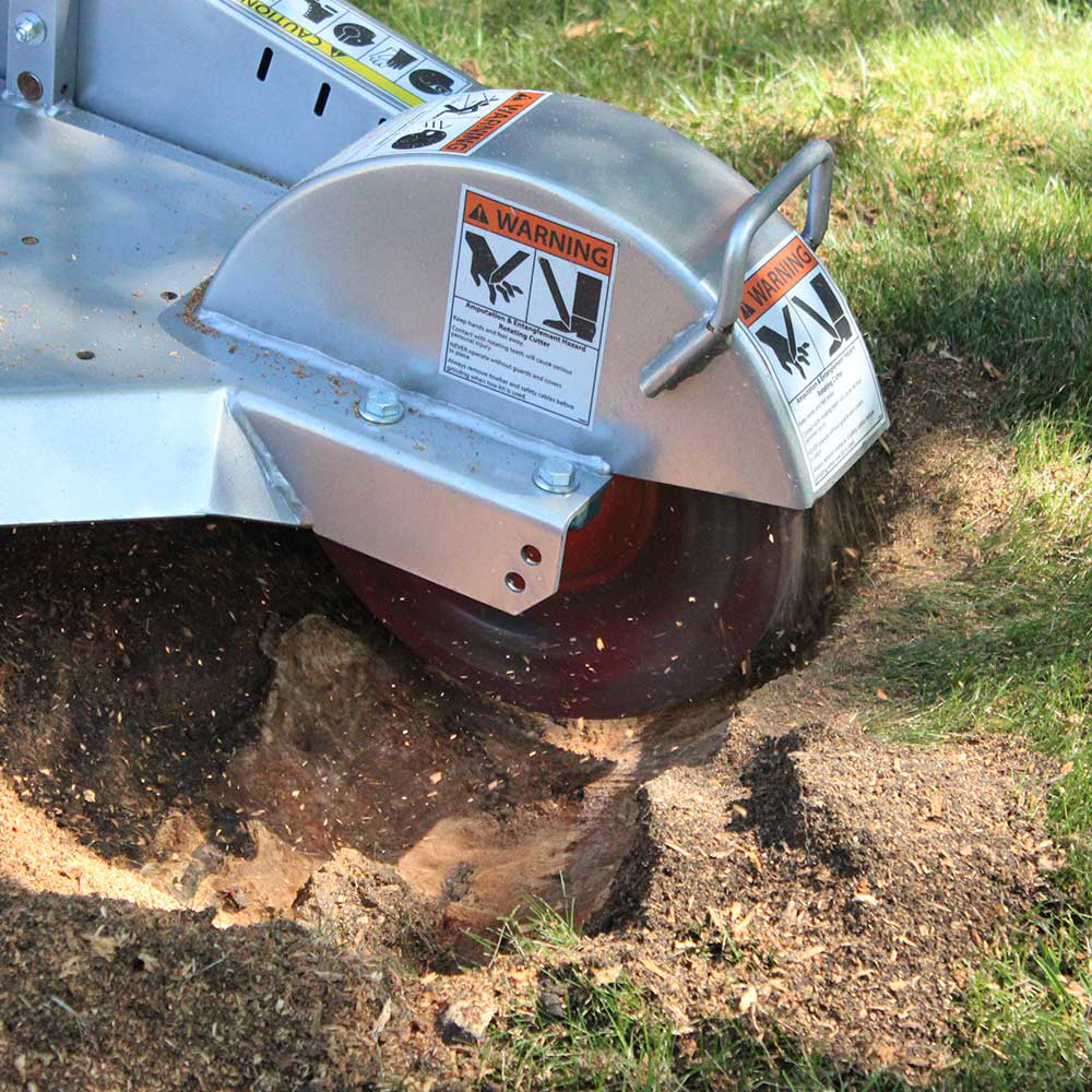 A close-up of a Dosko 337-13HC Stump Grinder's cutting wheel, which has a protective guard with a "WARNING" label, in the process of grinding a stump.