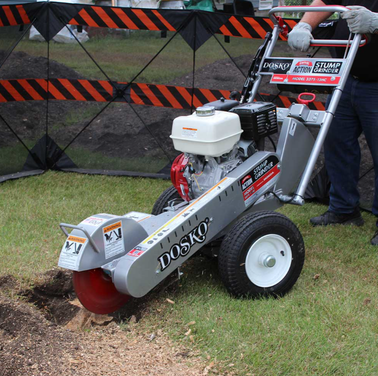 A Dosko 337S-13HC swivel stump grinder in action, with a person operating it in a grassy area with a warning fence in the background.