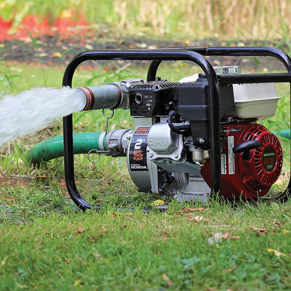 The Brave 2-inch semi-trash pump in action, actively pumping a stream of water through a hose onto a green, grassy lawn. The pump sits on the grass while operating.