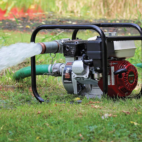 The Brave 2-inch semi-trash pump in action, actively pumping a stream of water through a hose onto a green, grassy lawn. The pump sits on the grass while operating.