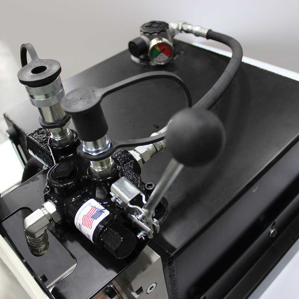 A close-up view of the manifold and control valve assembly of a Brave Hydra Buddy portable hydraulic power pack. The image shows the quick disconnect couplings, pressure gauge, and a control lever with a black ball handle. A small sticker with an American flag is visible on the manifold.