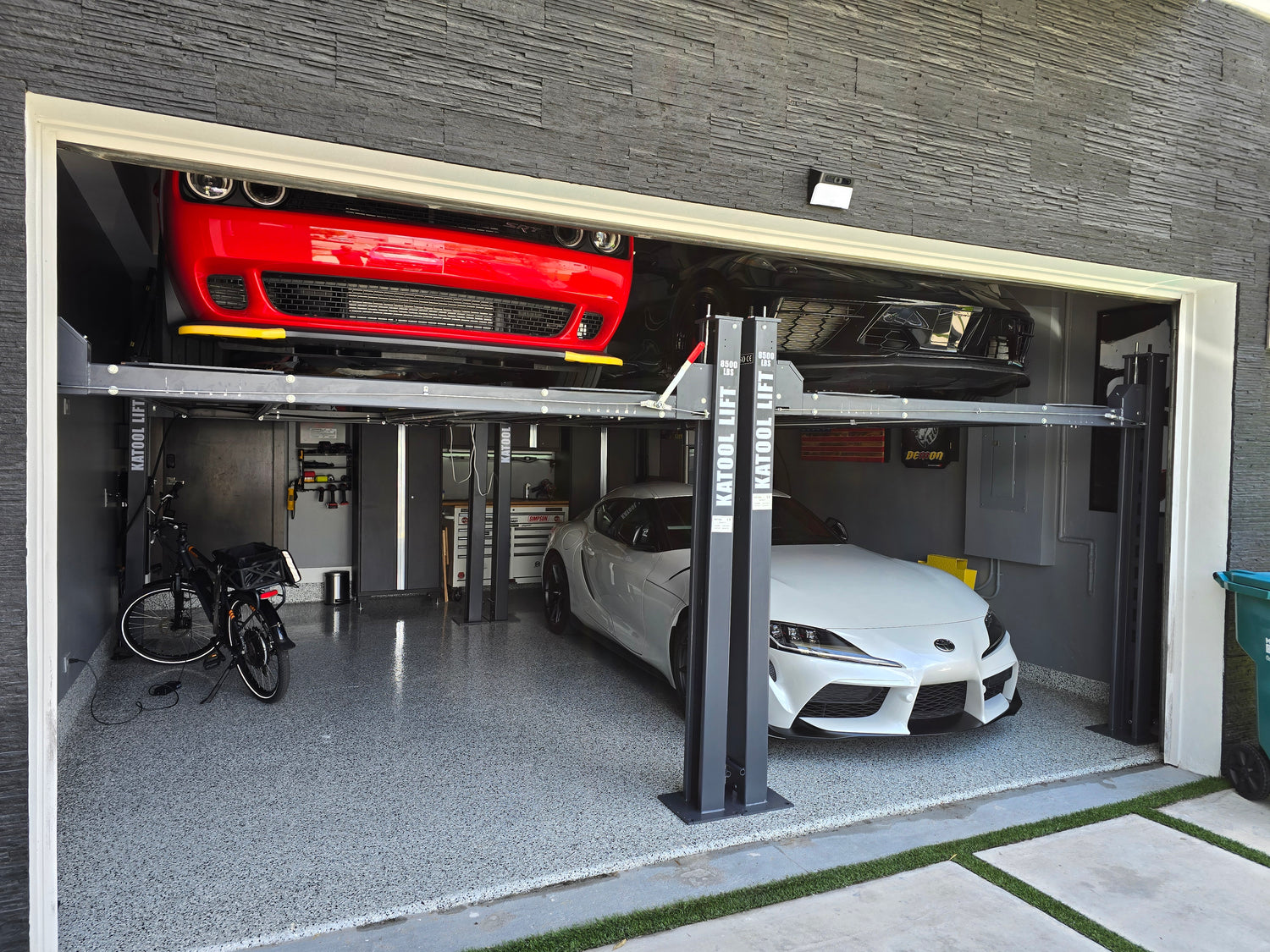Two cars, a red Dodge Challenger on the top platform and a white Toyota Supra below, are stored on a Katool car lift inside a modern garage