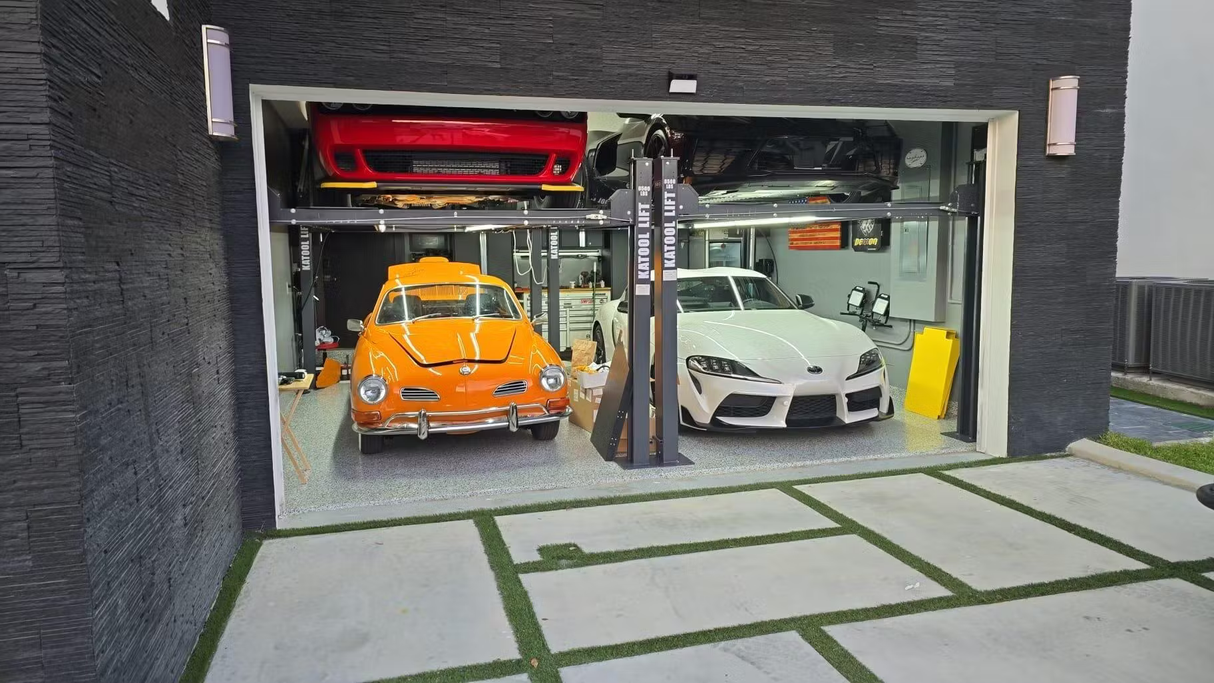 A red sports car on the top platform of the Katool car lift, with a white sports car below and an orange classic car parked nearby in a clean, modern garage