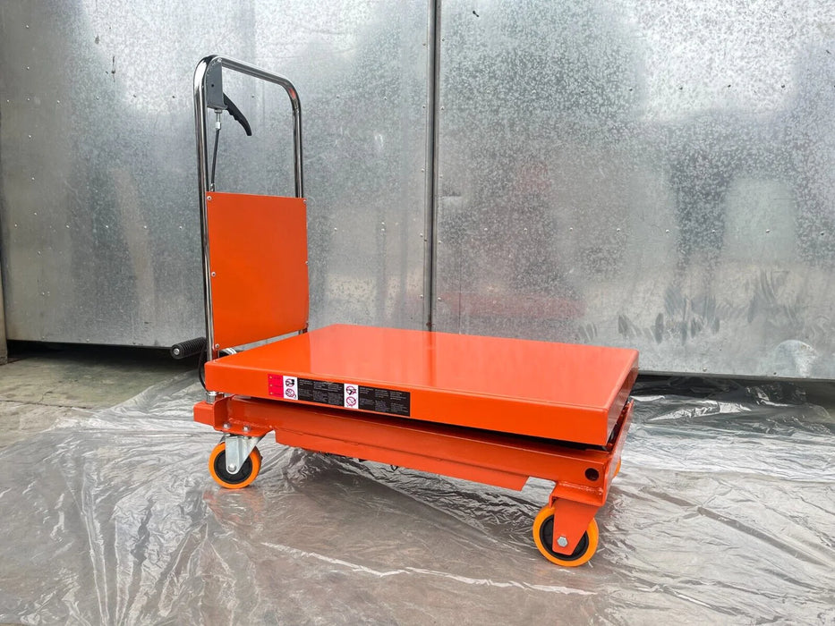A front view of the Katool Scissor Lift Table in a lowered position against a metal wall. The table has an orange finish, a black handle, and orange wheels with a locking mechanism.