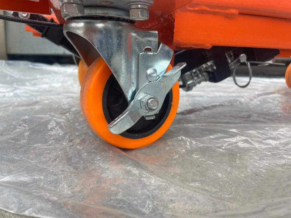 A close-up of a Katool Scissor Lift Table's orange swivel wheel, showing the metal caster and the foot-operated brake. The wheel is designed for secure and stable transportation.