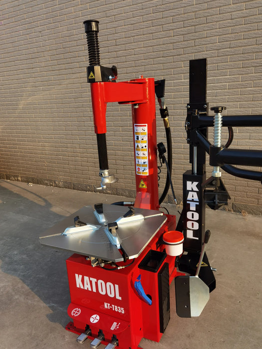 A full-body, three-quarter view of the red Katool KT-T835 tire changer, showcasing its rugged, compact design on a concrete surface. The machine's key components, including the assist arm and turntable, are clearly visible.