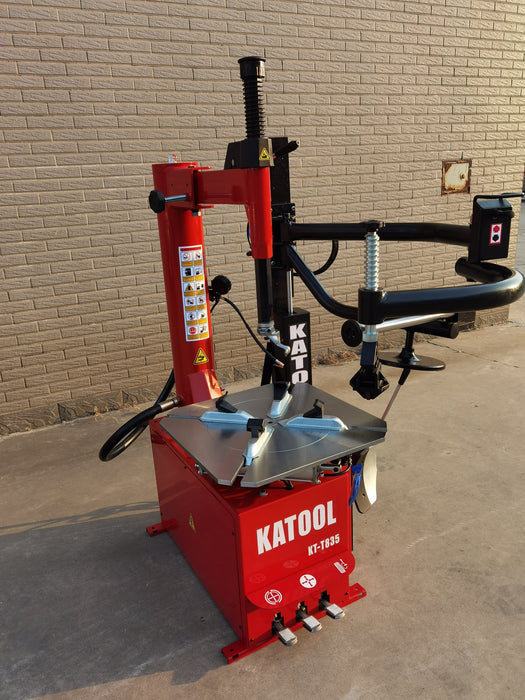 A full-body, three-quarter view of the red Katool KT-T835 tire changer. The machine is set against a brick wall, showcasing its closed-body design and the double-sided assist arm.