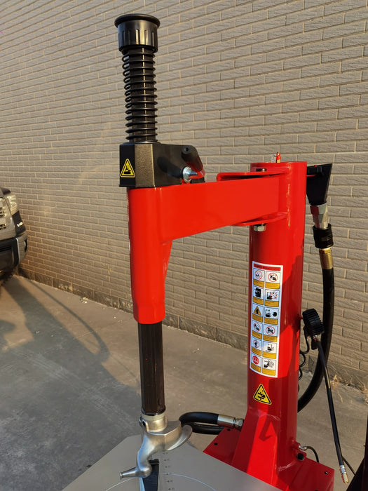 A side view of the red tower and a partial view of the turntable on the Katool KT-T835 tire changer. The durable construction and pneumatic components of the machine are visible against the brick wall background.