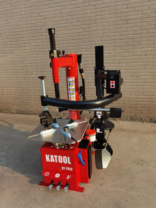 A full-body, three-quarter view of the Katool KT-T835 tire changer. The machine is shown against a brick wall, showcasing its professional design with a red body, assist arm, and a prominent bead breaker on the side.
