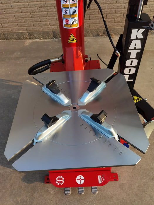 A top-down view of the silver turntable on the Katool KT-T835 tire changer, showing the four clamping jaws in a wide-open position. The red control pedal for the bead breaker is visible at the bottom of the machine.