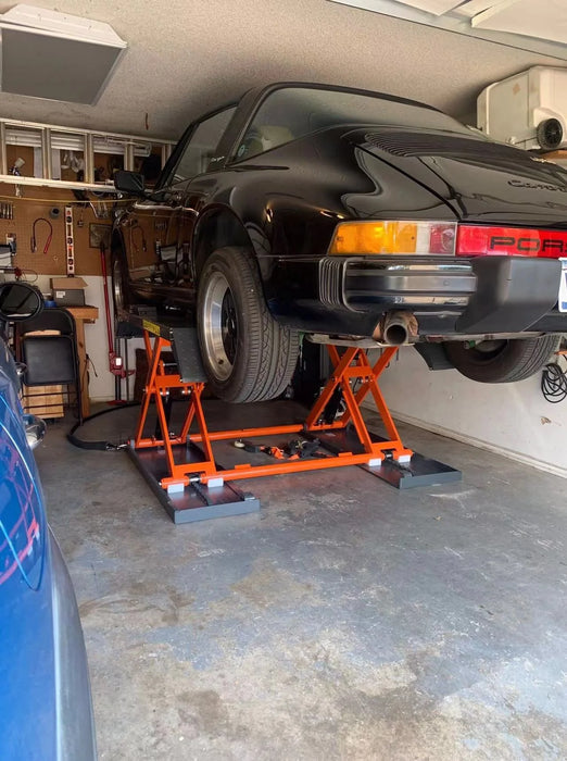 A wide-angle shot of a black classic Porsche raised on the orange Katool KT-X80-ELECTRIC scissor lift in a well-lit garage, with tools and equipment on the wall.