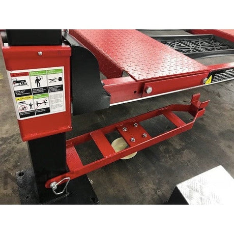A close-up view of the caster kit for the NOS9000XLT lift, which is a red metal frame with white wheels, positioned next to the lift's platform and a warning label.