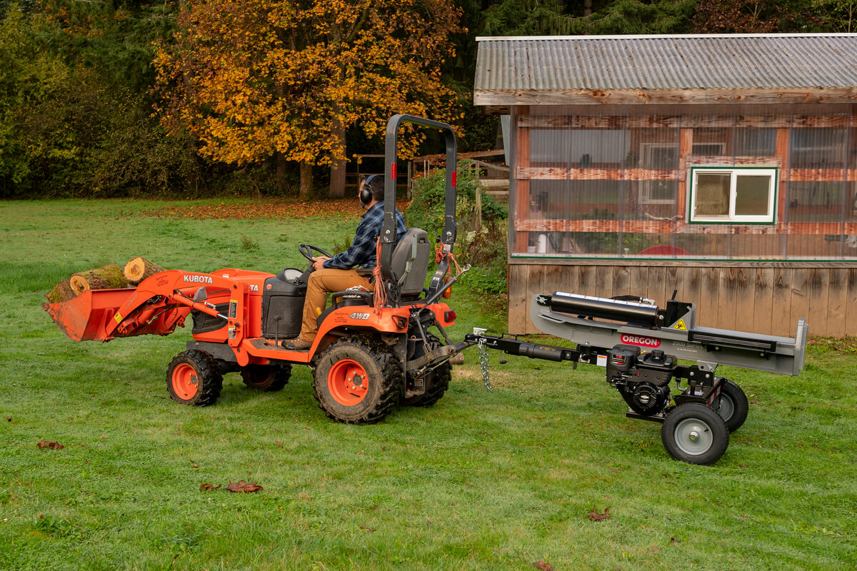 The Oregon 2500 Series 25-ton log splitter is being towed by a small orange Kubota tractor in a grassy field with a small building in the background.