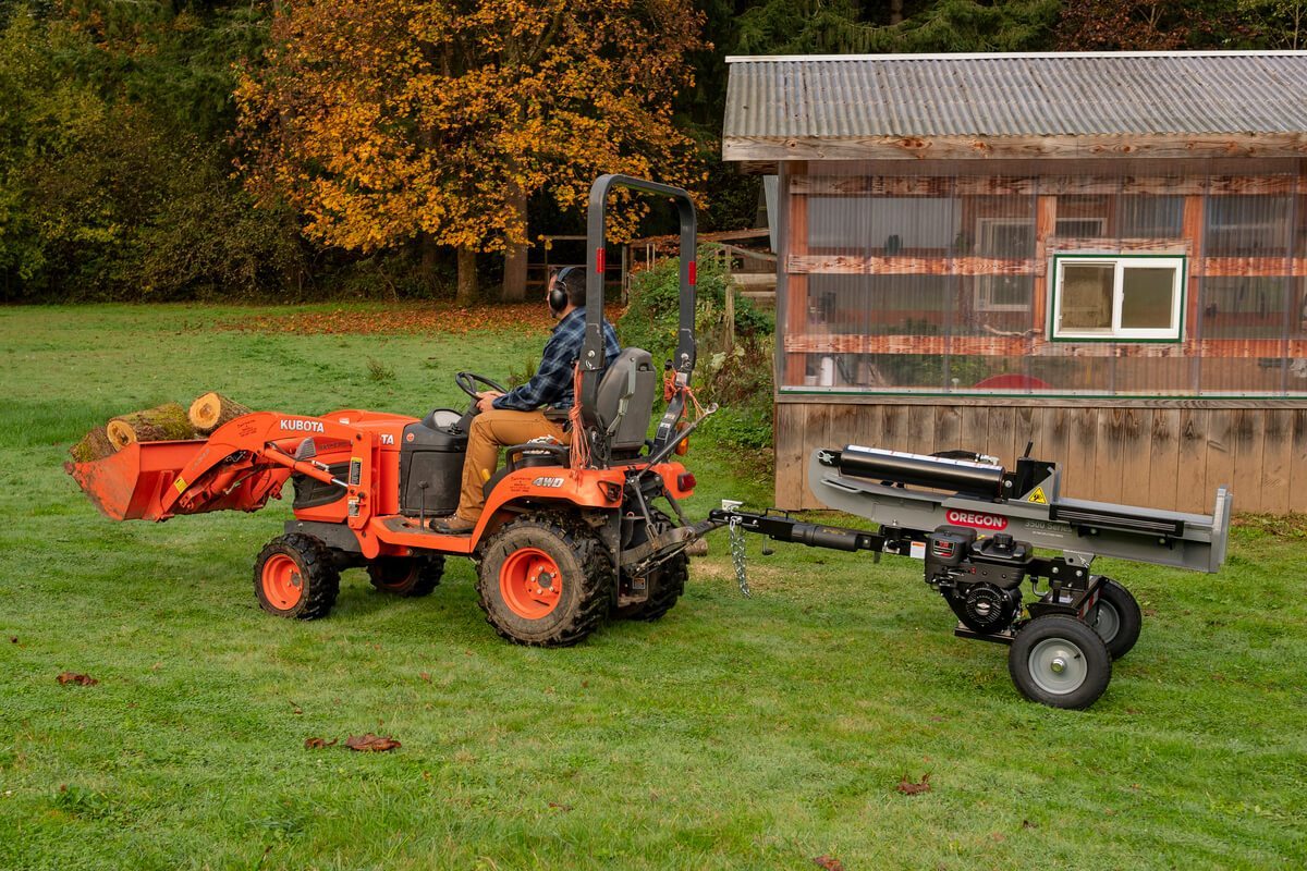 The Oregon 3500 Series 35-ton log splitter is being towed by a small orange Kubota tractor in a grassy field with a small building in the background.