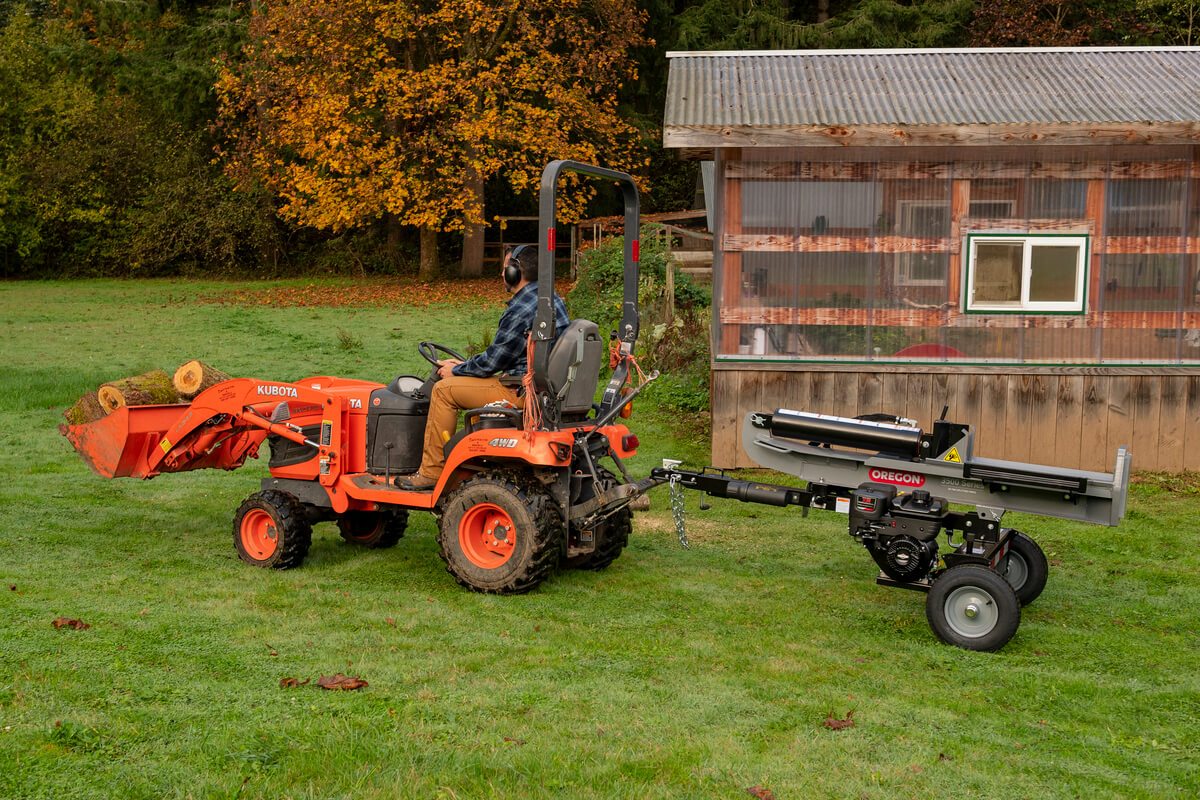 The Oregon 3000 Series 30-ton log splitter is being towed by a small orange Kubota tractor in a grassy field with a small building in the background.